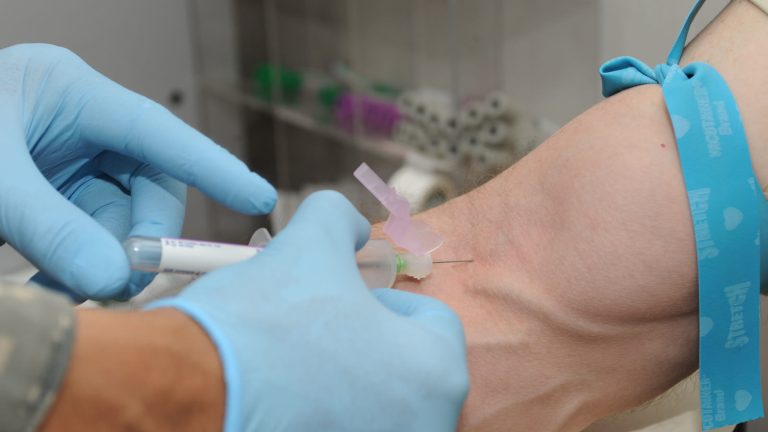 Vårdpersonal med blå engångshandskar tar ett blodprov ur armvecket på en patient