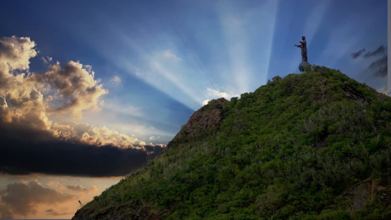 Cristo Rei-statyn står på en grönklädd kulle mot blå himmel med solstrålar och moln, öster om Dili i Timor-Leste