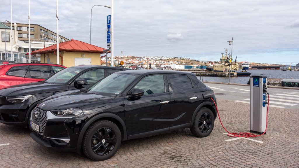 Svart elbil som laddas vid en laddstolpe på en kullerstensparkering vid hamnen i Lysekil, med fler bilar bredvid